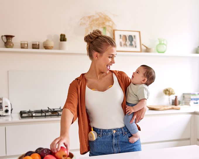 A mom holds her baby and has a snack while wearing her Medela Freestyle™ Hands-free double electric breast pump.
