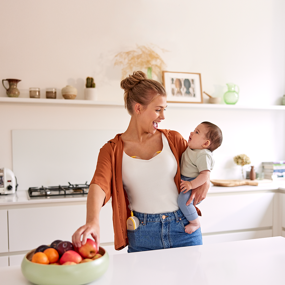 Mom holding baby and grabbing a snack while wearing her Medela Freestyle™ Hands-free double electric wearable breast pump