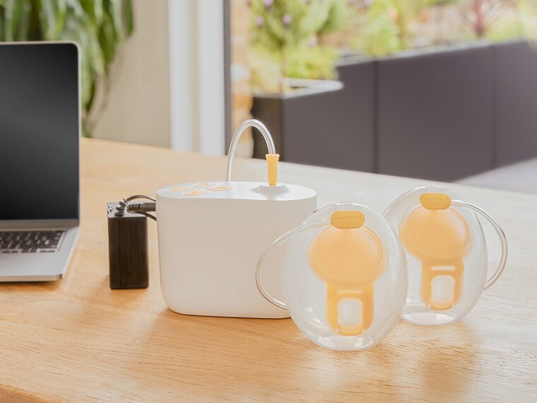 Medela Pump In Style® Hands-free double electric breast pump sitting on office desk with the pump, Hands-free collection cups and battery pack charger