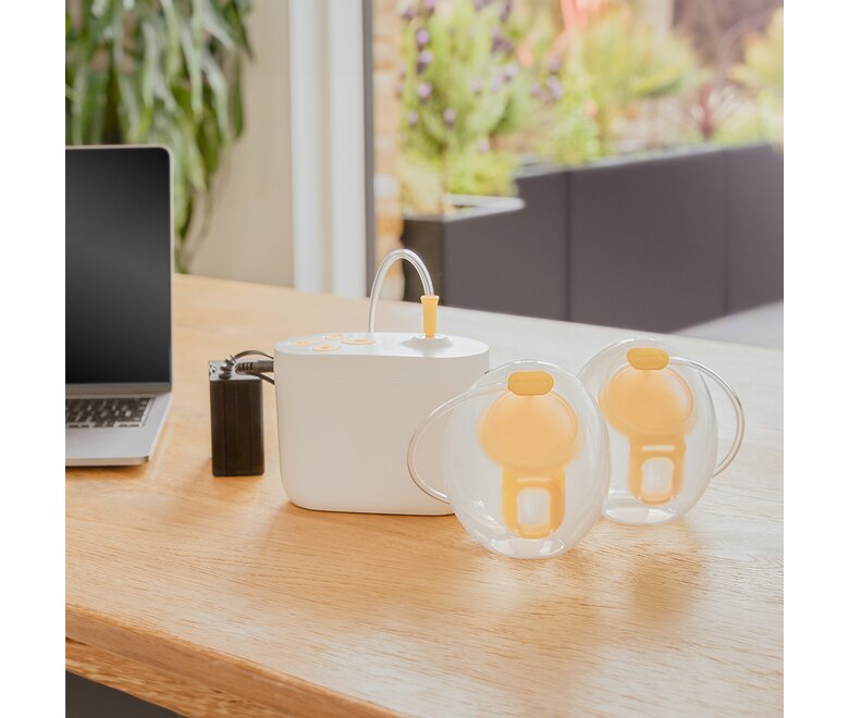Medela Pump In Style® Hands-free double electric breast pump sitting on office desk with the pump, Hands-free collection cups and battery pack charger