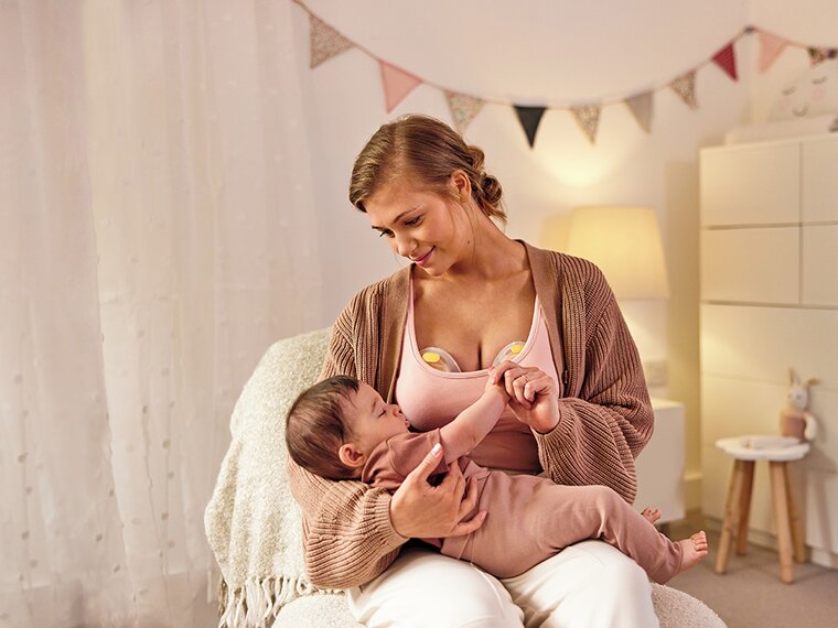 Eine Mutter trägt eine elektrische Medela Swing Maxi™ Doppelmilchpumpe, während sie ihr Baby im Kinderzimmer hält.
