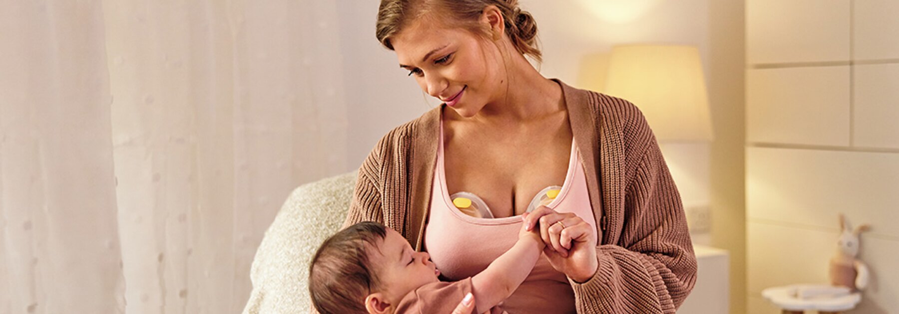 Eine Mutter trägt eine elektrische Medela Swing Maxi™ Doppelmilchpumpe, während sie ihr Baby im Kinderzimmer hält.