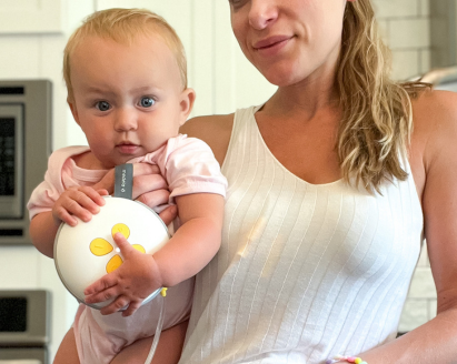 Woman standing and holding her baby while she wears Swing Maxi Hands-free breast pump.