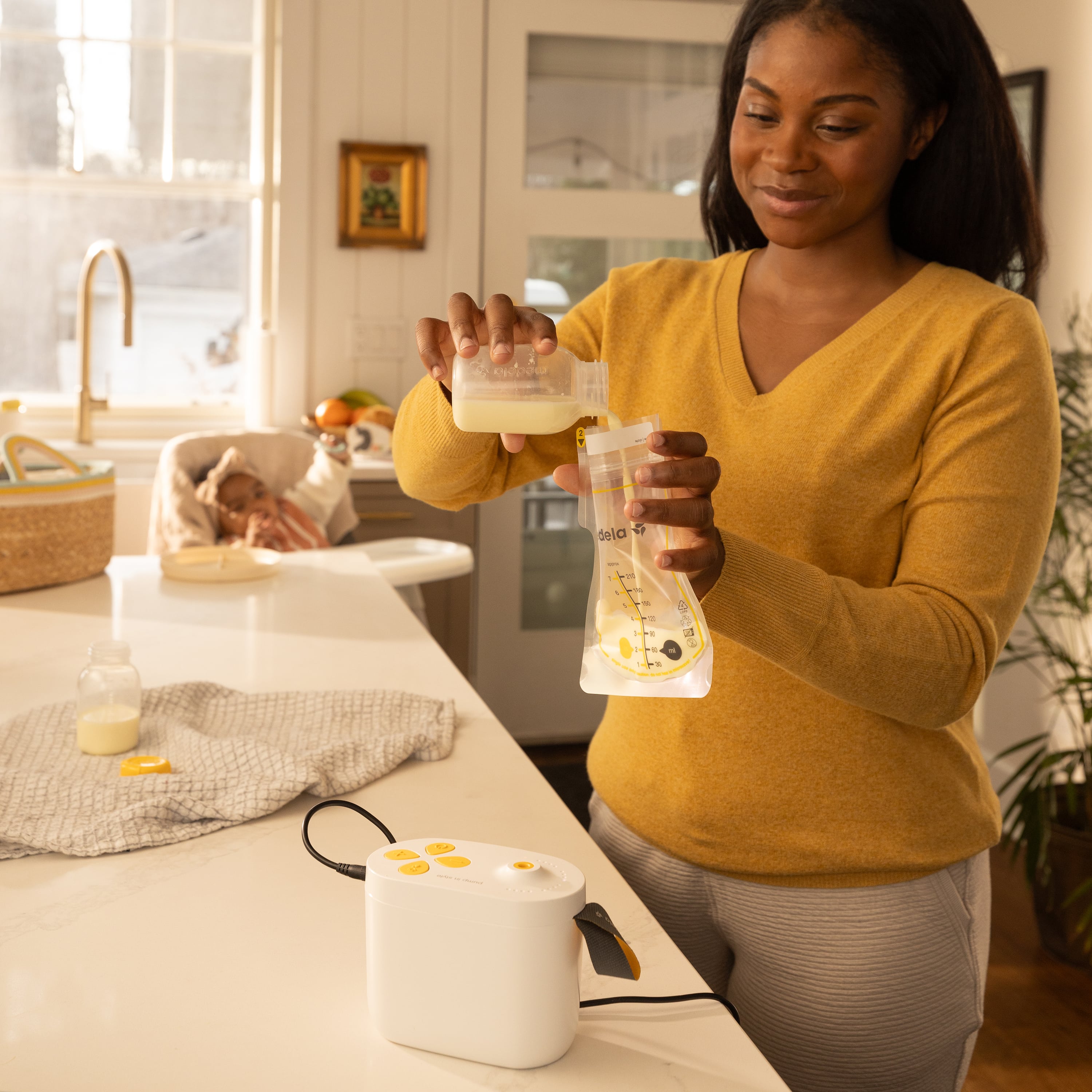 Woman in kitchen pouring breast milk expressed with Pump In Style Pro breast pump into Easy Pour Breast Milk Storage Bag.  Baby is sitting in high chair watching mom.