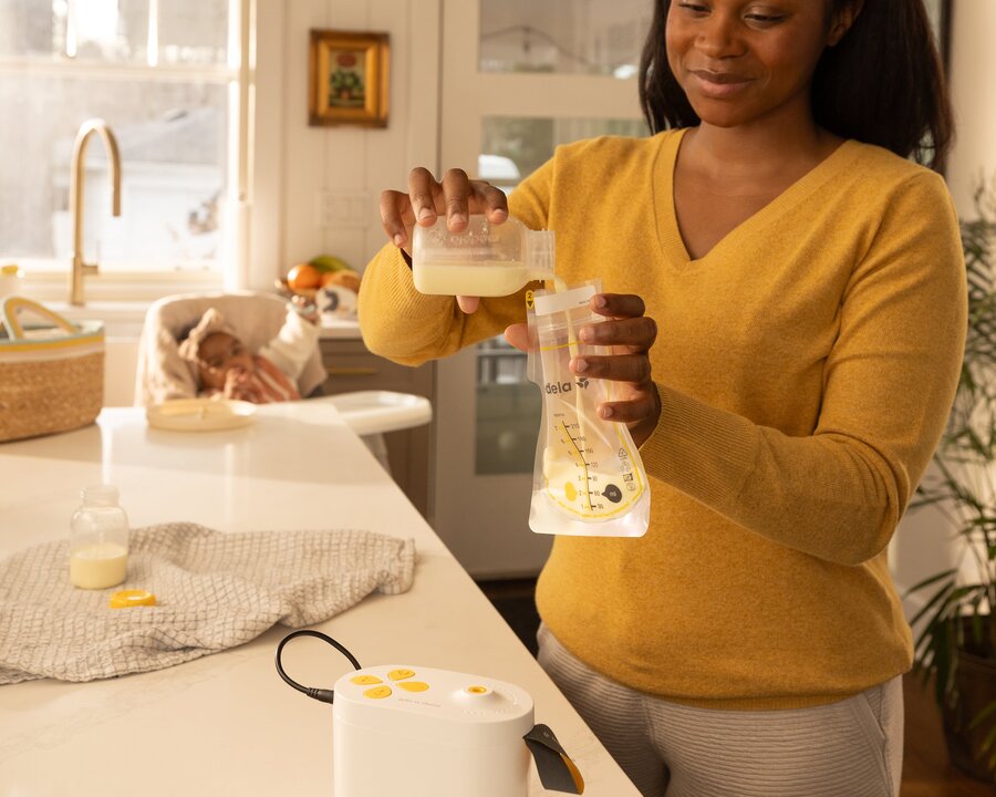 Pump In Style Pro breast pump and Easy Pour Breast Milk Storage Bags. Woman in kitchen pouring breast milk expressed with Pump In Style Pro breast pump into Easy Pour Breast Milk Storage Bag. Baby is sitting in high chair watching mom.