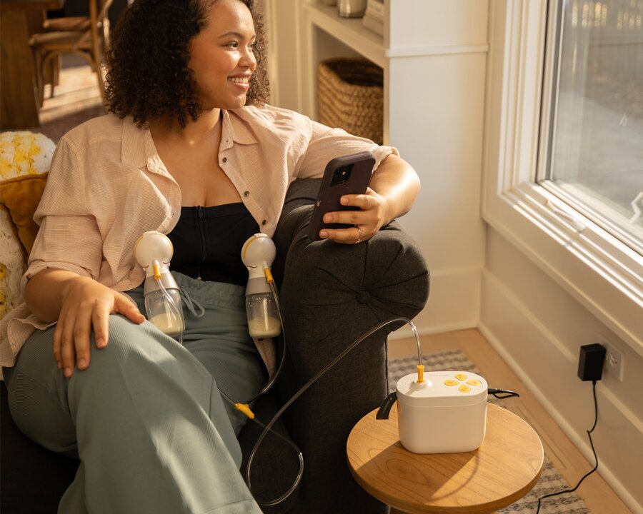 Woman sitting on couch, holding her phone, and looking out the window while using her Pump In Style Pro breast pump.