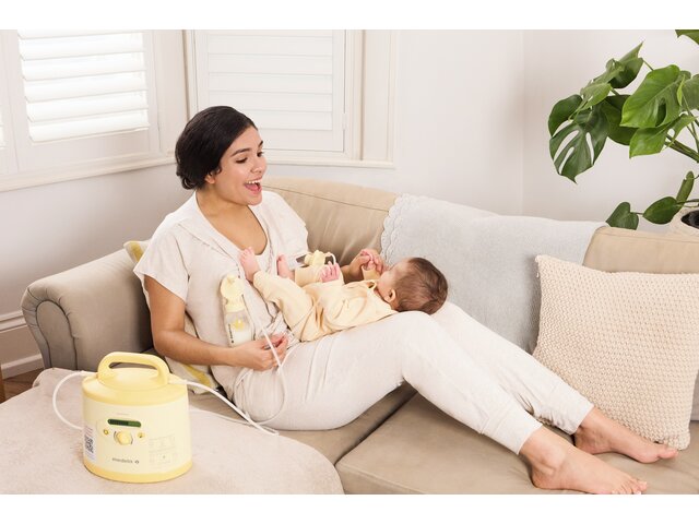 Una mamá juega con su bebé en el regazo mientras está tumbada en el sofá y se extrae leche con su sacaleches Medela Symphony PLUS® alquilado.