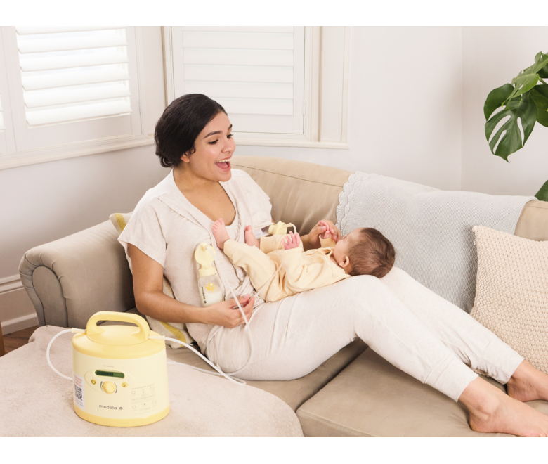 Eine Mutter spielt mit ihrem Baby auf dem Schoß, während sie auf der Couch liegt und mit ihrer gemieteten Medela Symphony PLUS® Milchpumpe abpumpt.