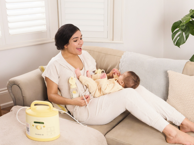 Una mamma gioca con il suo bambino in grembo mentre è sdraiata sul divano e pompa con il suo tiralatte Medela Symphony PLUS® noleggiato.