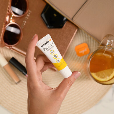 A person holds a 7 g tube of Medela Purelan™ lanolin cream in their hand over sunglasses, make-up and tea for on the go.