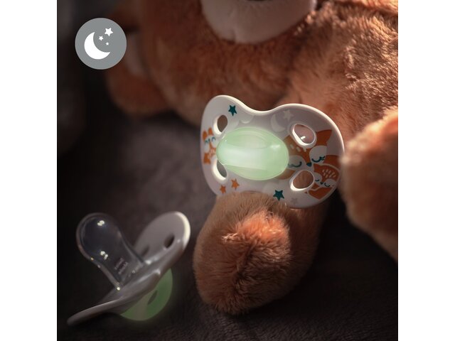 The Medela NIGHT & NIGHT dummies with forest animal designs lie on a bed with a toy bear. Includes a “glow in the dark” symbol.