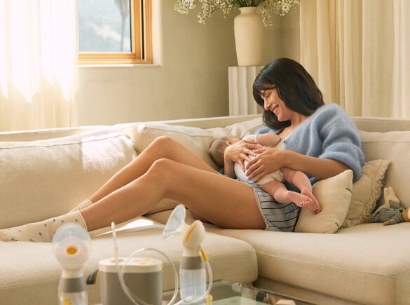 Eine Person hält ein Baby auf einem Sofa, während eine Medela Pump in Style Milchpumpe auf einem nahegelegenen Tisch steht.