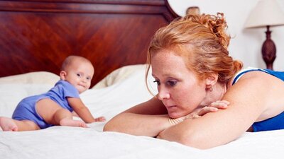 A mom leans against her bed and sighs as her baby crawls and looks back at her.