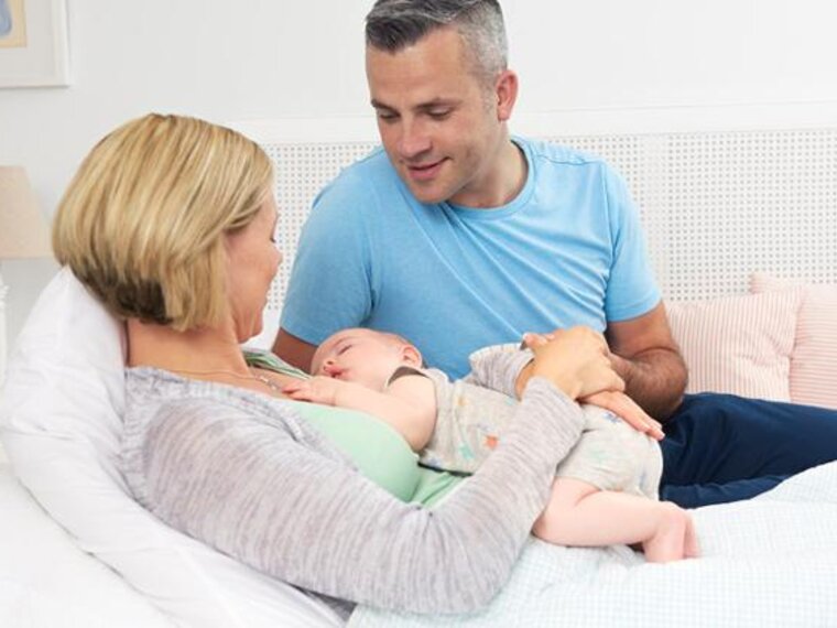 A mom lies on the bed while the baby sleeps on her chest, the dad sits on the bed next to them.