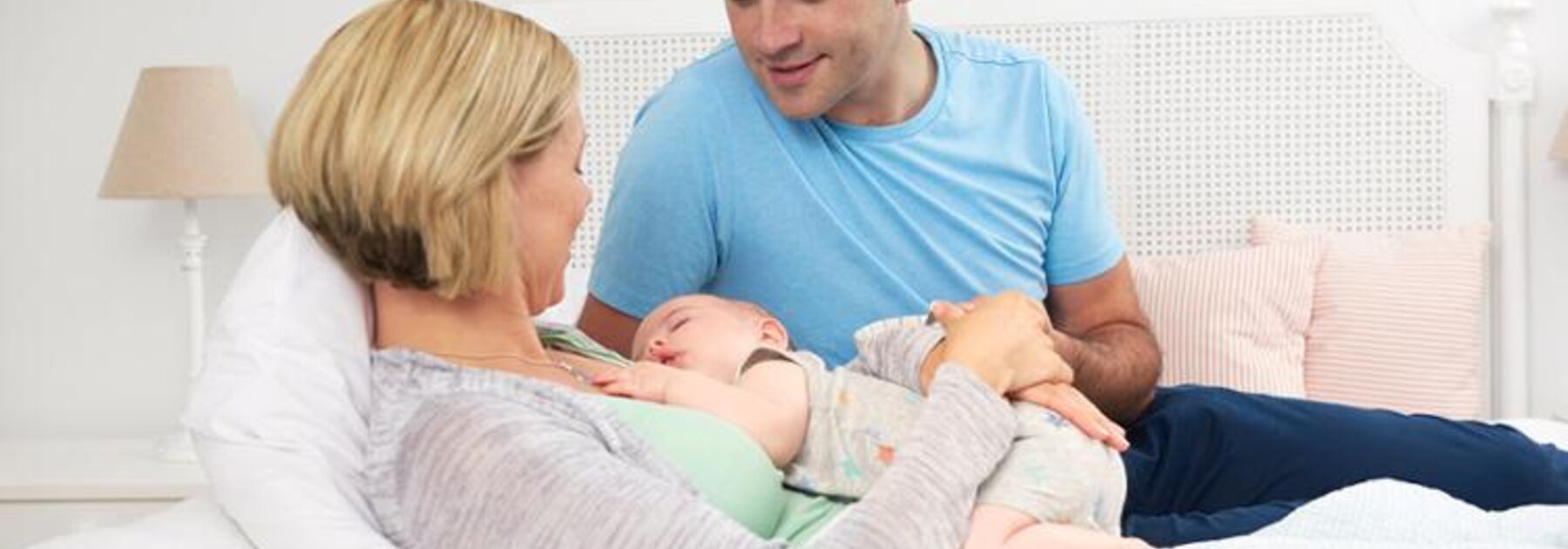 A mom lies on the bed while the baby sleeps on her chest, the dad sits on the bed next to them.