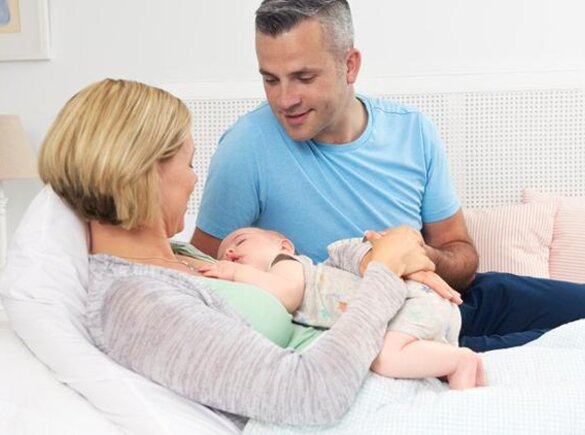 A mom lies on the bed while the baby sleeps on her chest, the dad sits on the bed next to them.