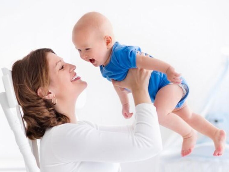 A mother in a rocking chair plays with her baby and laughingly lifts him up and down.
