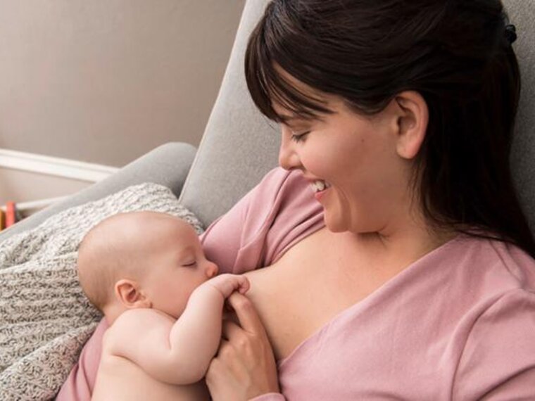 A mother smiles and looks down at her baby as they nurse, sitting at home in the living room.