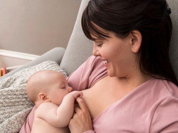 A mother smiles and looks down at her baby as they nurse, sitting at home in the living room.