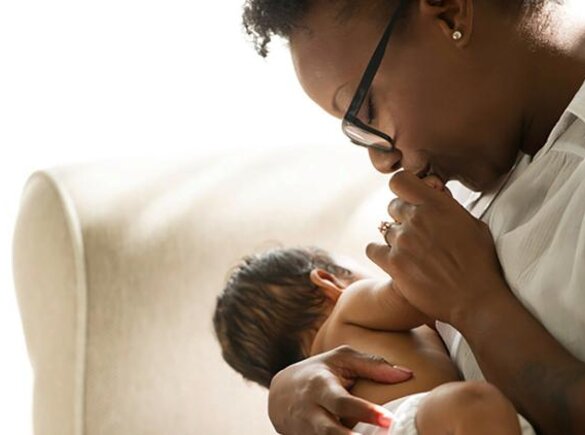 A baby is nursing while its mother kisses its hand as they sit in a chair at home.
