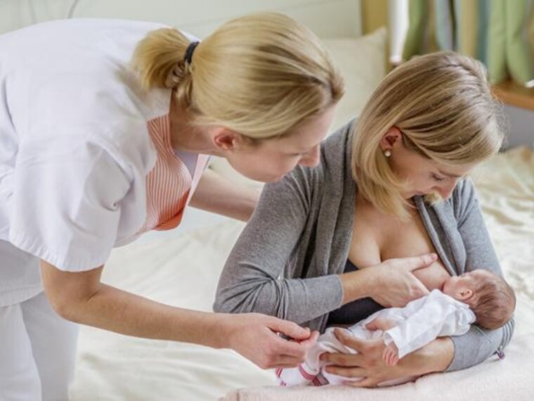 A mom breastfeeds her newborn baby while sitting at the end of the bed.