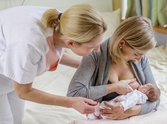 A mom breastfeeds her newborn baby while sitting at the end of the bed.