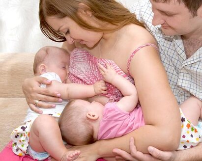 Una mamma allatta i suoi gemelli in tandem, mentre il papà si siede accanto a loro e sostiene uno dei bambini.