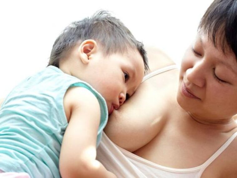 A mother is lying on her left side while her son is breastfeeding on her right side and resting quietly against her.