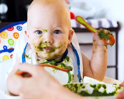 A parent feeds his baby in a highchair with green vegetables as baby food.
