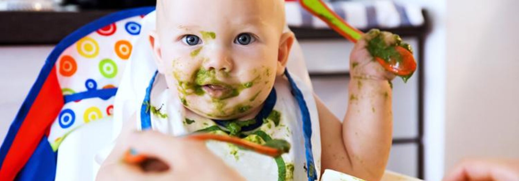 A parent feeds his baby in a highchair with green vegetables as baby food.