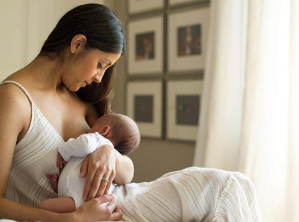 A mother holds her newborn baby while it is breastfed, sitting on the bed near the window at home.
