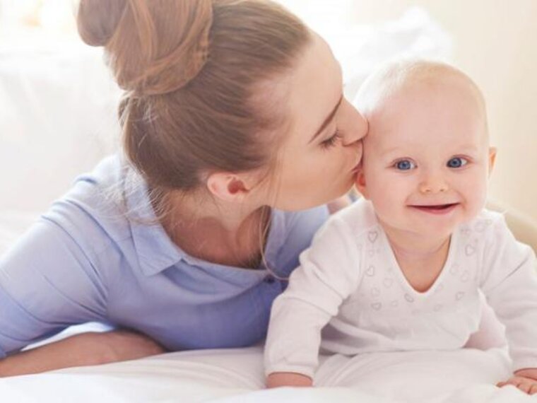 A baby crawls around on the bed while its mother lies next to it and gives it a kiss on the side of the head.