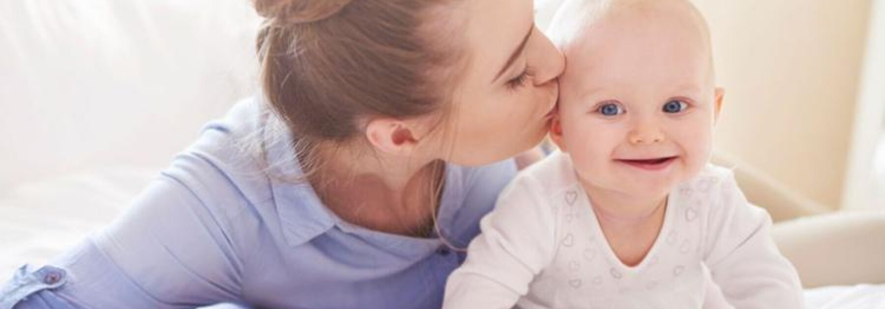 A baby crawls around on the bed while its mother lies next to it and gives it a kiss on the side of the head.