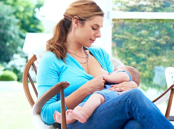 A mom breastfeeds her baby while sitting in a rocking chair by the window.