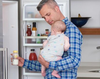 Een vader met zijn baby in zijn armen neemt een fles moedermelk uit de koelkast om op te warmen voor een voeding.