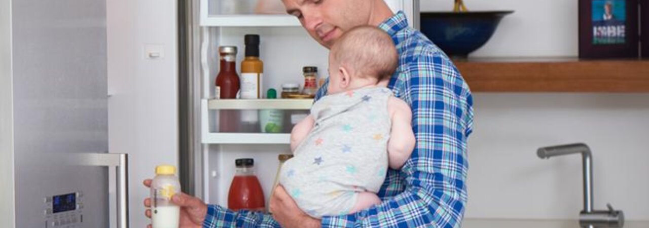 Een vader met zijn baby in zijn armen neemt een fles moedermelk uit de koelkast om op te warmen voor een voeding.