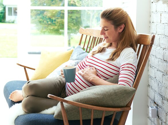 Eine schwangere Frau sitzt draußen auf der Veranda mit einem Getränk in der einen und der anderen Hand auf ihrem schwangeren Bauch.
