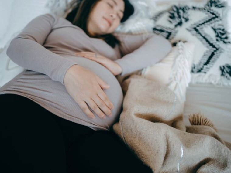 A pregnant woman lies asleep on her side in bed, holding her pregnant belly.