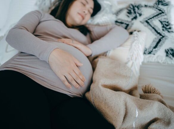 Een zwangere vrouw ligt op haar zij in bed te slapen en houdt haar zwangere buik vast.