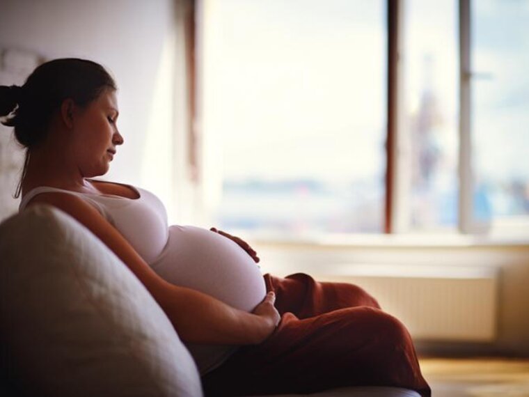 Een zwangere vrouw zit op een bank in een schemerige kamer met haar handen op haar babybump en kijkt tevreden naar beneden.