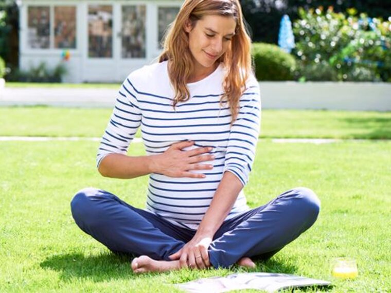 A pregnant woman sitting outside on the grass looking at a book.