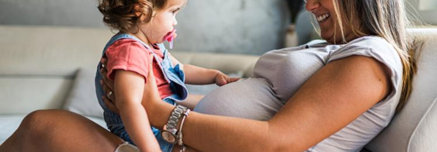 A pregnant woman sits on a couch with her toddler daughter on her lap, facing each other.