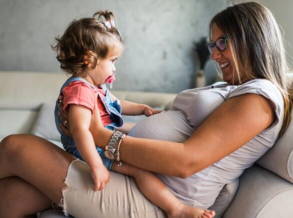 A pregnant woman sits on a couch with her toddler daughter on her lap, facing each other.