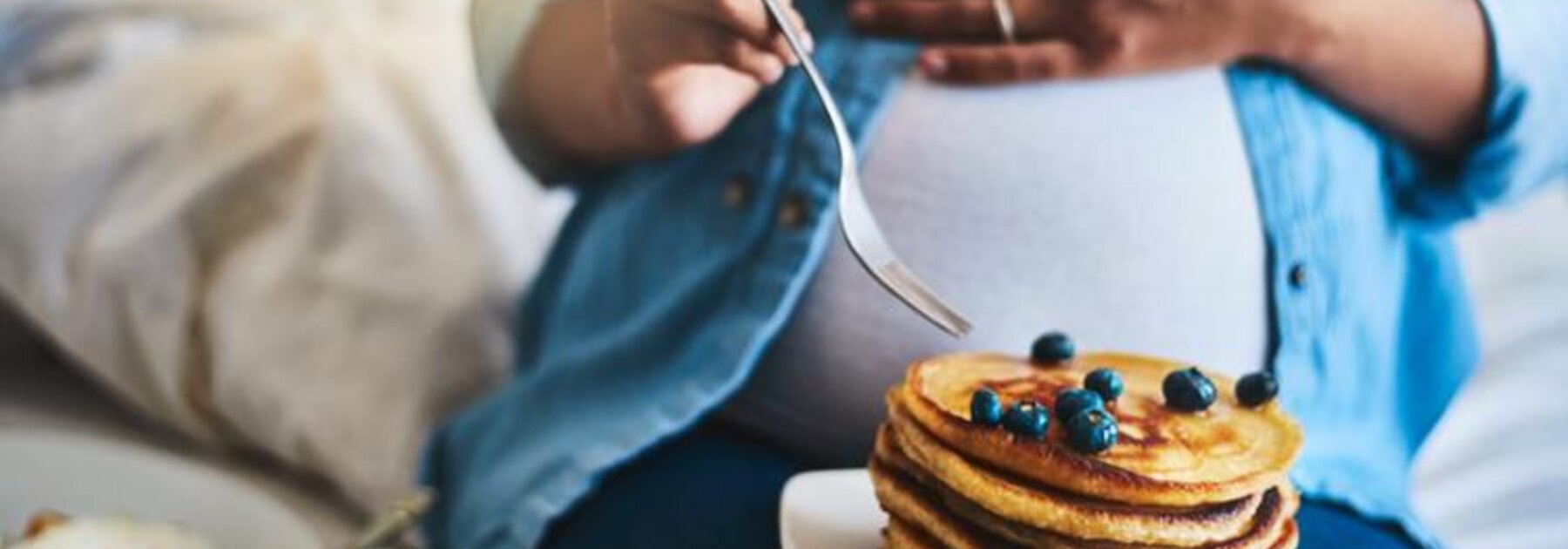 Een zwangere vrouw die haar buik vasthoudt terwijl ze pannenkoeken en fruit eet, zit op een bank.