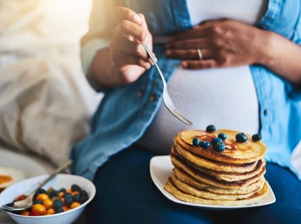 Een zwangere vrouw die haar buik vasthoudt terwijl ze pannenkoeken en fruit eet, zit op een bank.