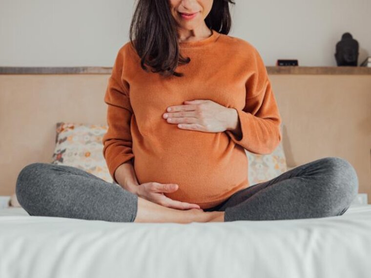 A pregnant mother sits at the end of a bed with her legs drawn up and her hands on her stomach.