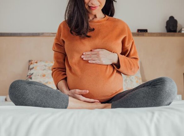 A pregnant mother sits at the end of a bed with her legs drawn up and her hands on her stomach.
