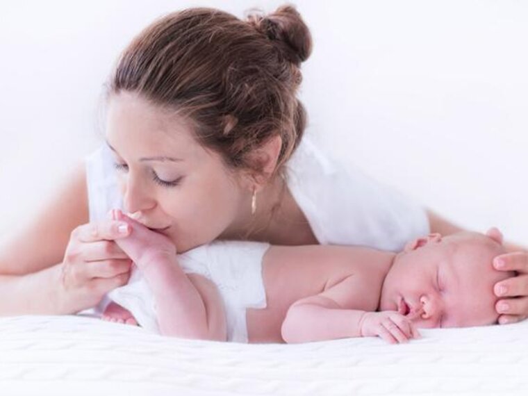 A baby sleeps on its tummy while its mother gives it a gentle kiss on the bottom of its foot and places her hand on its head.