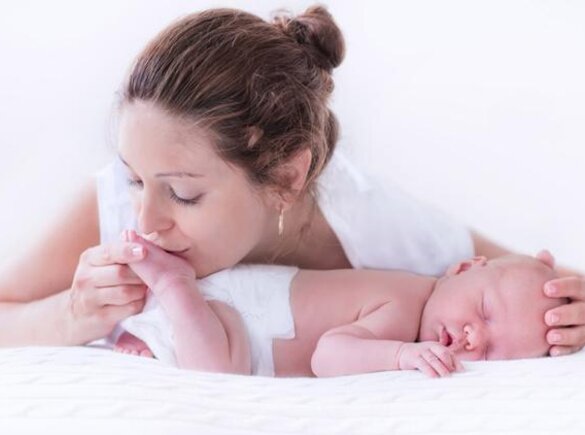 A baby sleeps on its tummy while its mother gives it a gentle kiss on the bottom of its foot and places her hand on its head.