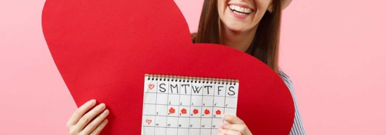 Eine glückliche Frau hält ihren Schwangerschaftskalender hoch, hinter dem sich ein großes rotes Papierherz befindet.